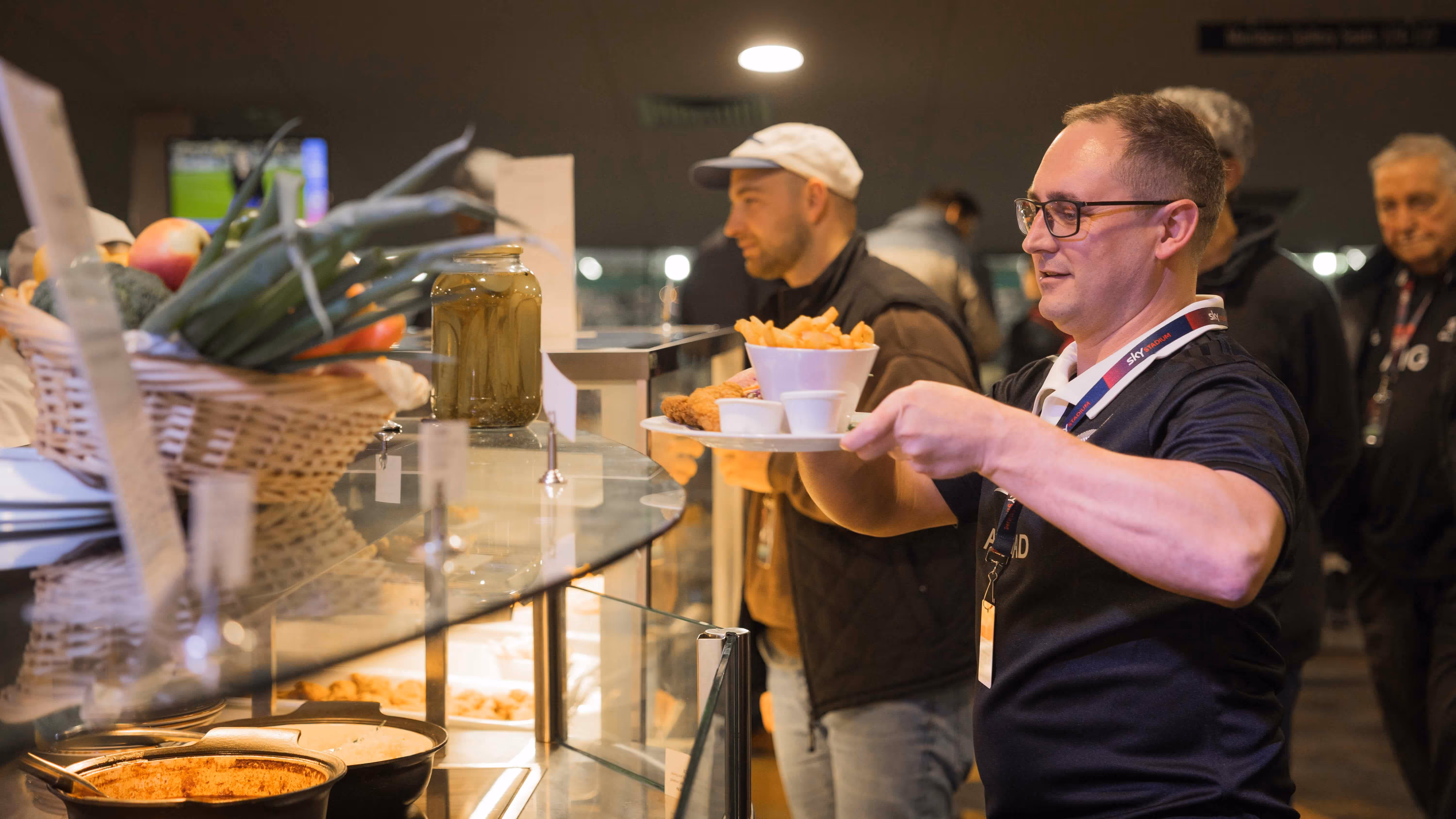 Man serving a plate of fries, fried chicken, and dipping sauces at a food counter with other people waiting in line.
