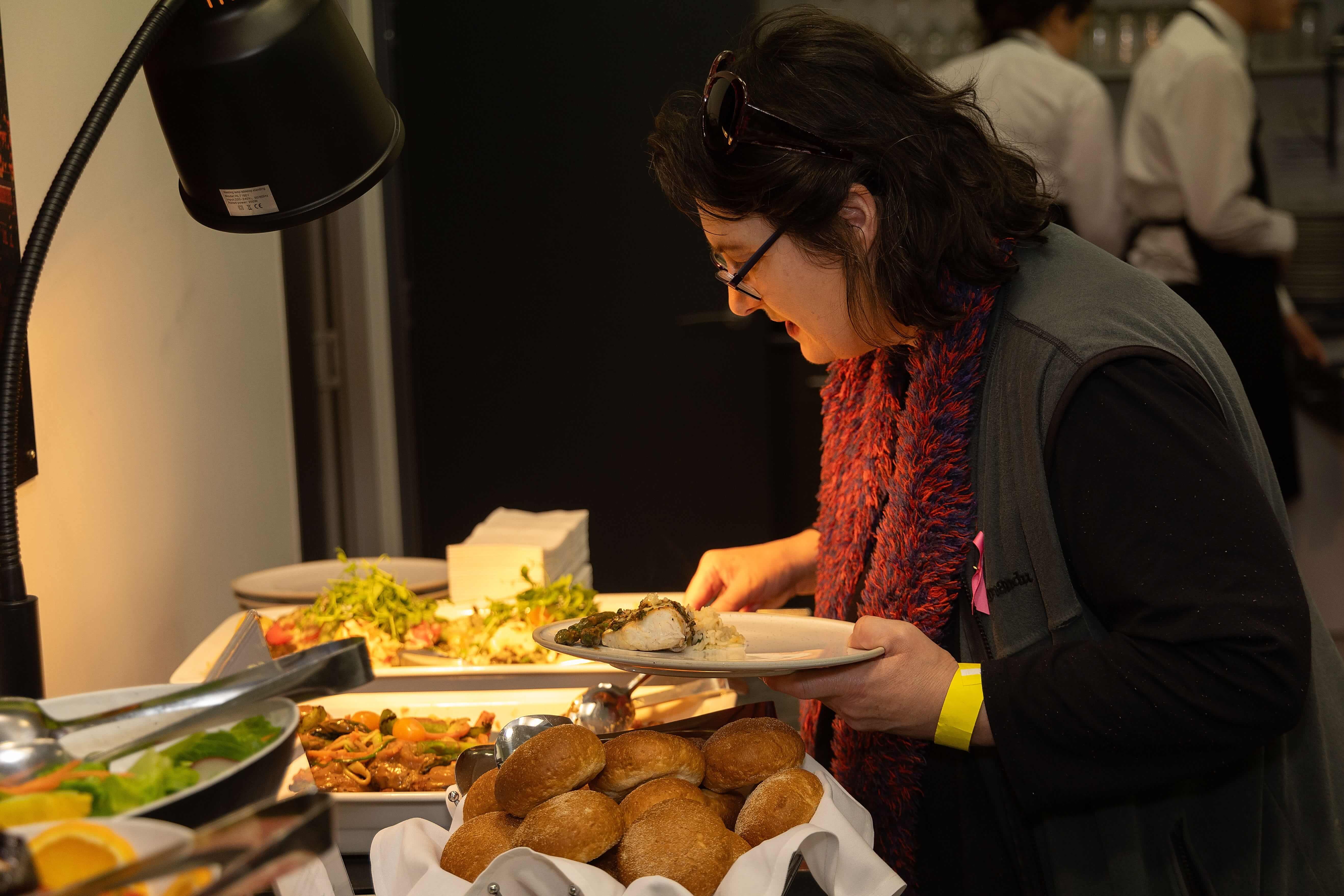 Woman in glasses and a red scarf serving food onto a plate at a buffet table with bread rolls and various dishes.