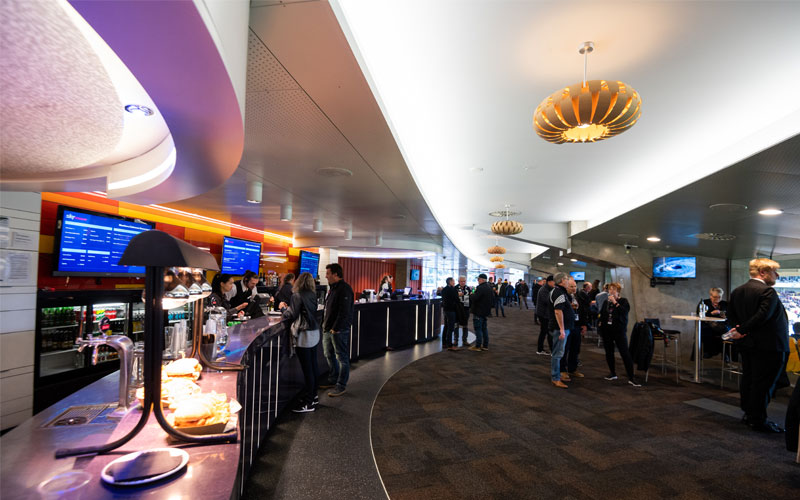 People socializing and ordering drinks at a modern bar inside a spacious clubroom with curved ceiling lights.