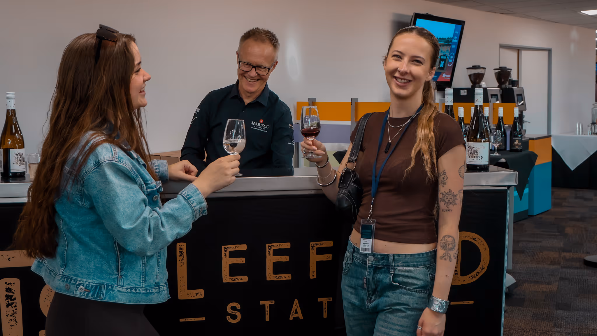 Two women holding wine glasses and smiling near a counter with a man behind it, bottles of wine displayed on the counter in a tasting room.