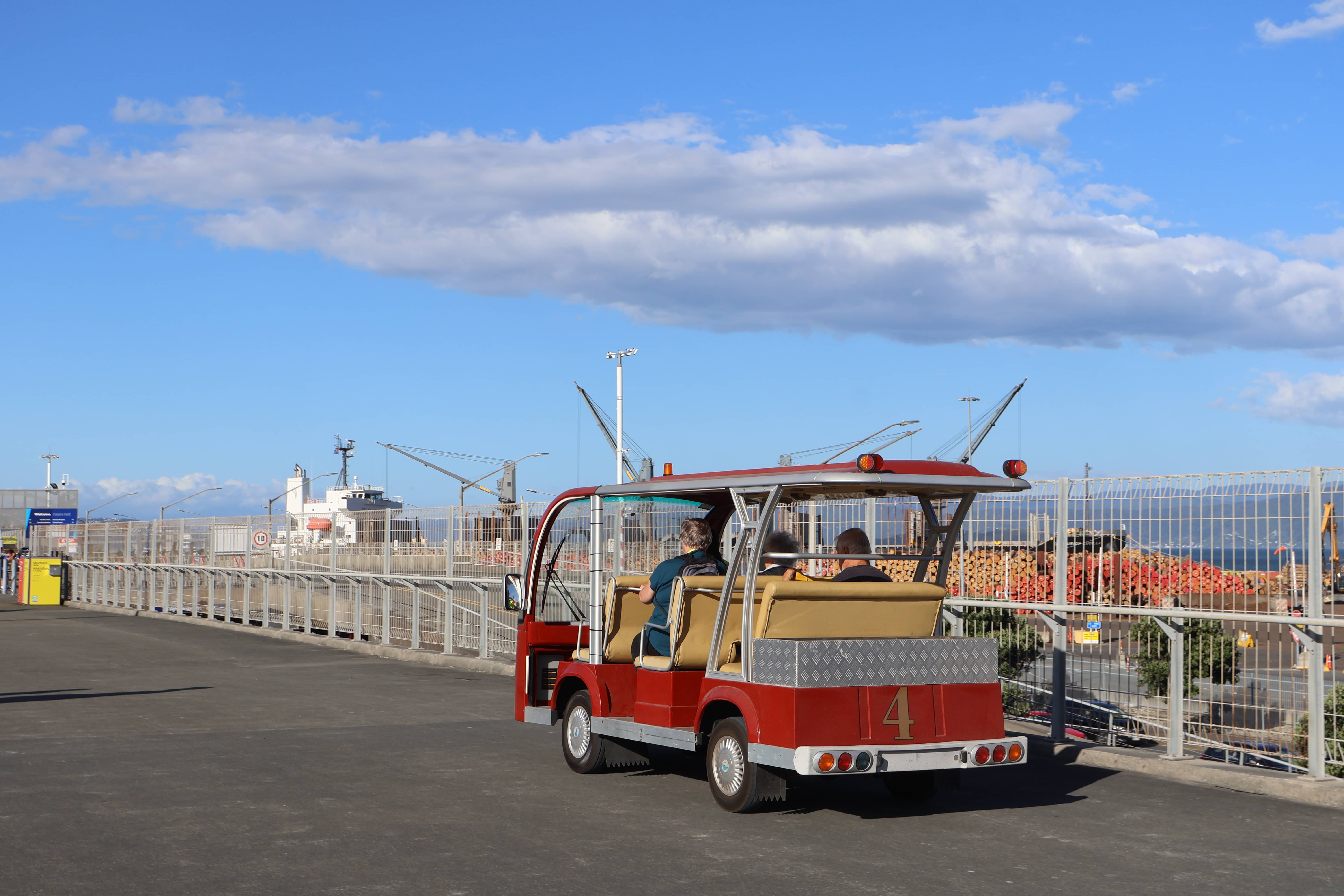 Small red open-sided utility vehicle with passengers driving on a paved area near a fenced port with cranes and stacked timber.