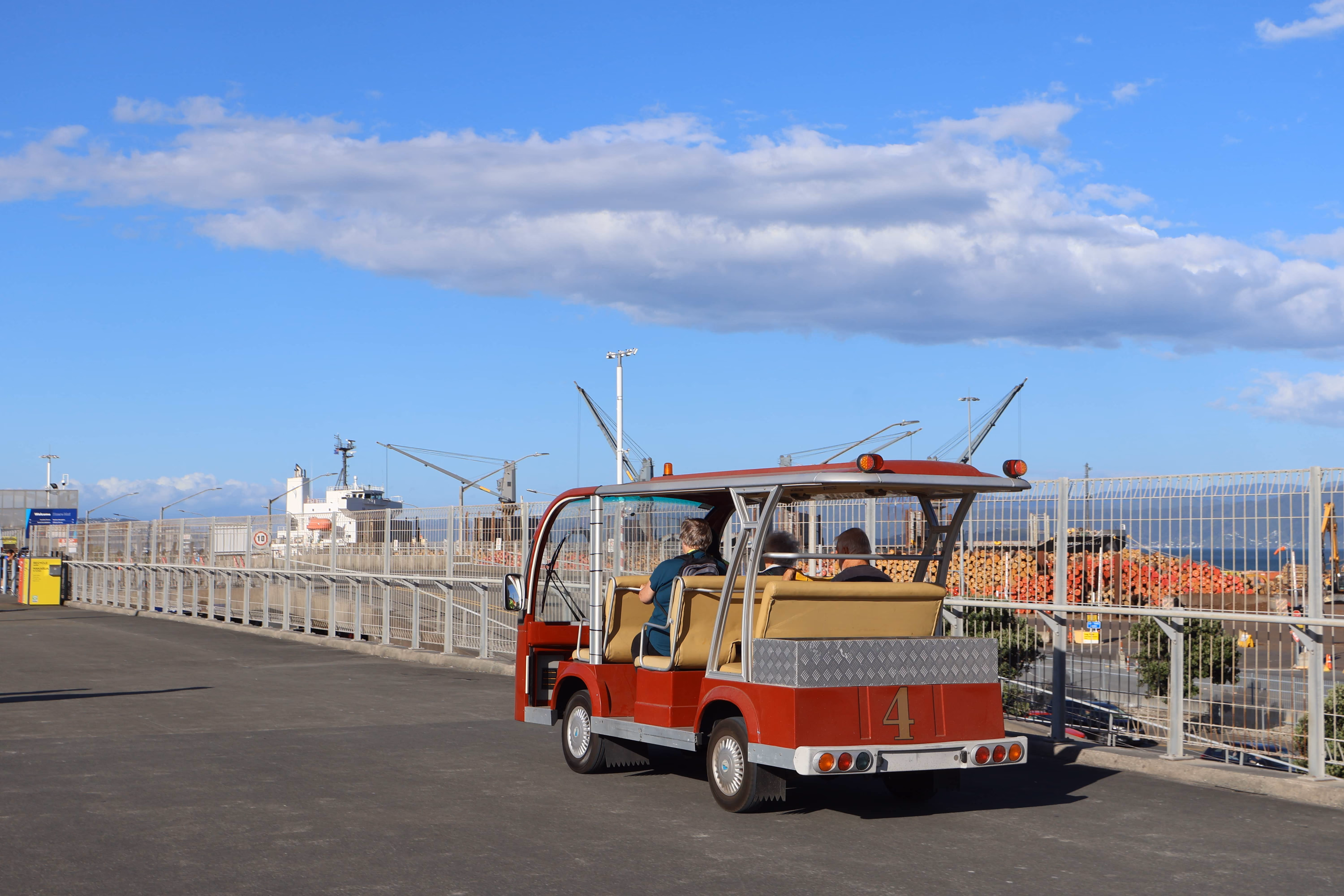 Small red open-sided utility vehicle with passengers driving on a paved area near a fenced port with cranes and stacked timber.