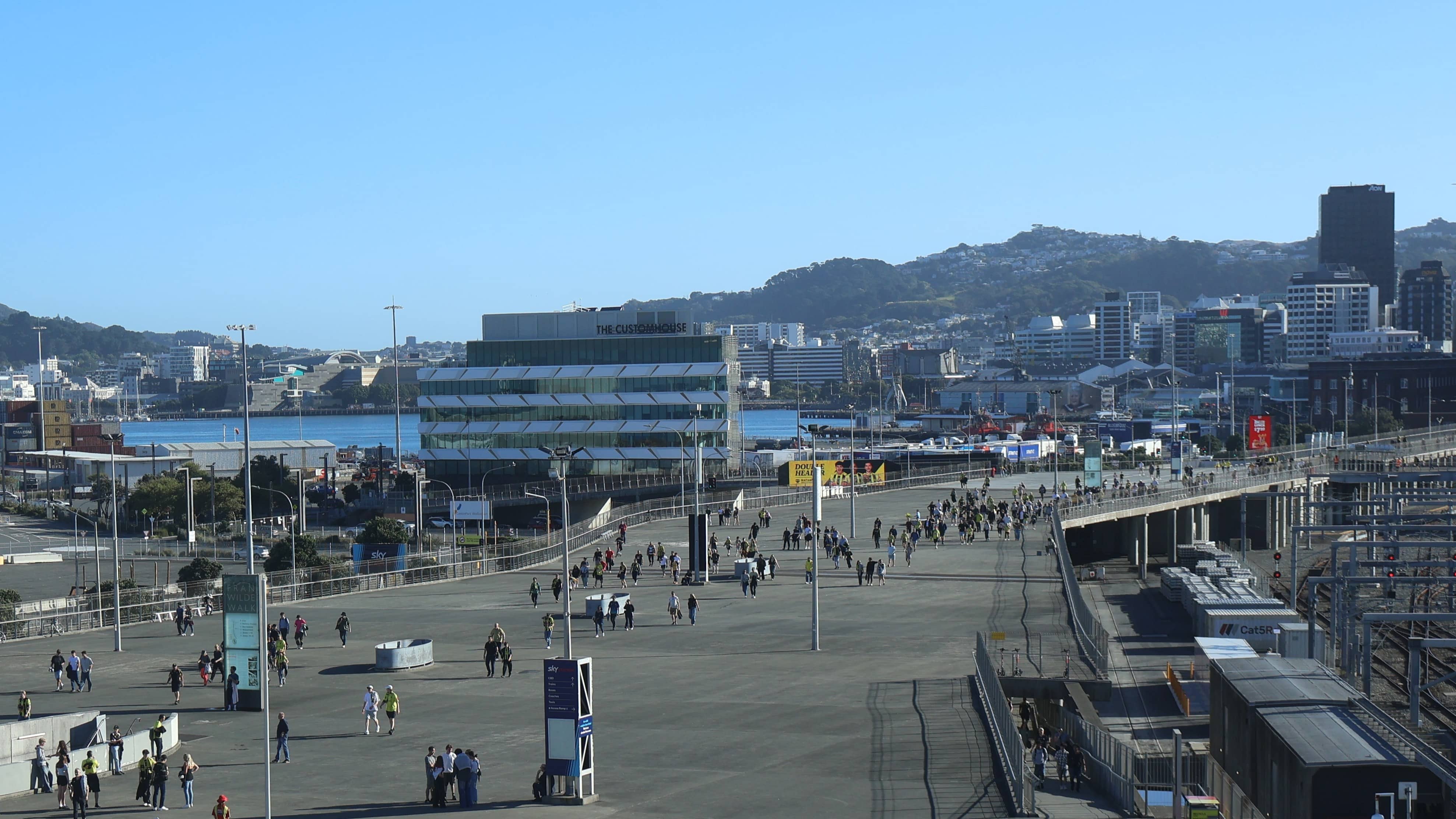 Wide view of a waterfront promenade with people walking, modern buildings, and hills in the background under a clear blue sky.