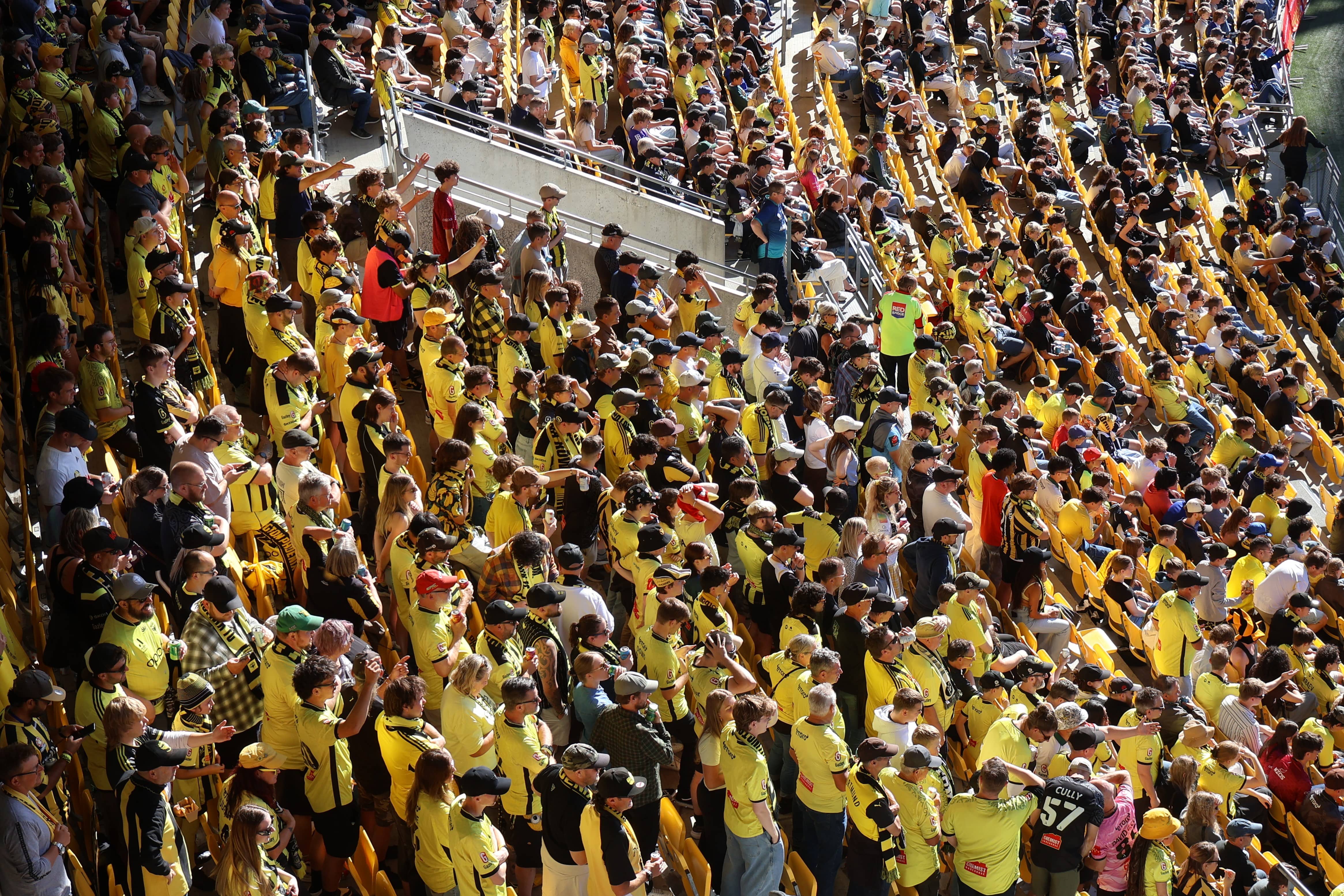 Crowd of sports fans wearing yellow and black seated and standing in stadium bleachers during a daytime event.