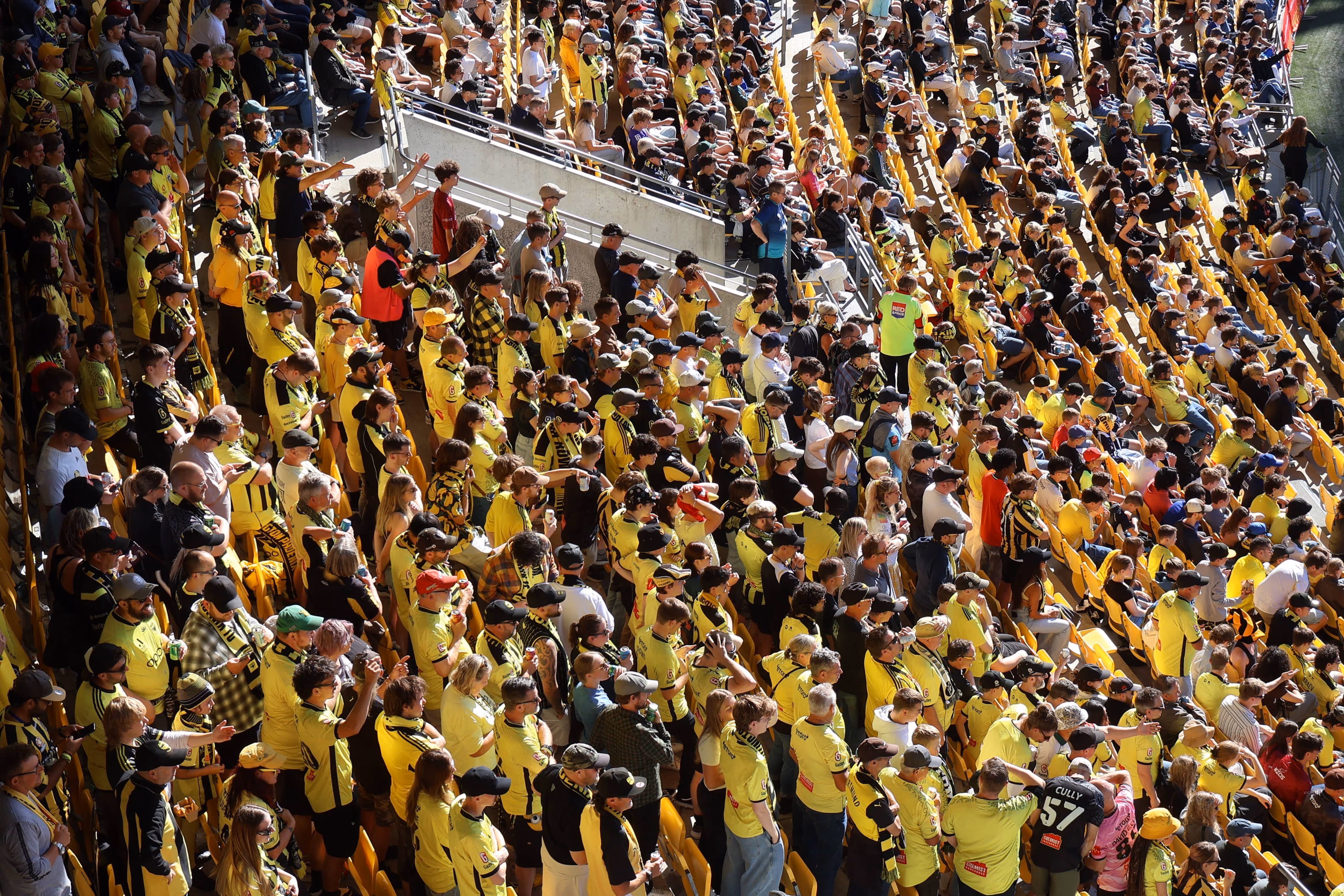 Crowd of sports fans wearing yellow and black seated and standing in stadium bleachers during a daytime event.