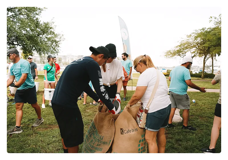 Group of people outdoors participating in a community cleanup, filling large burlap sacks labeled 'Cafe Bras' with collected trash.