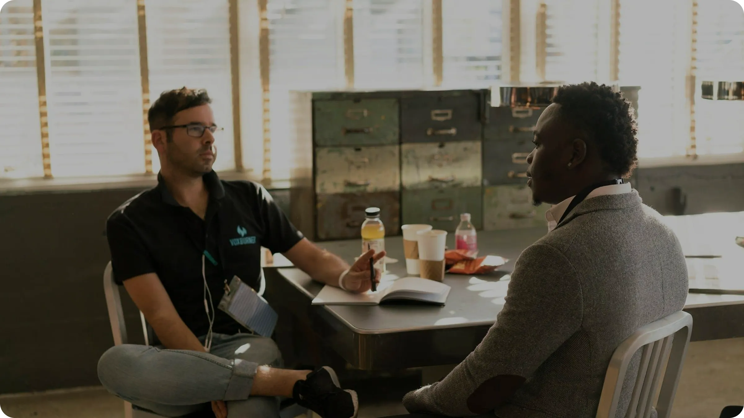 Two men sitting at a table engaged in conversation in a sunlit room with large windows and metal filing cabinets.