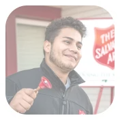 Smiling young man holding a small red shovel in front of a Salvation Army sign.