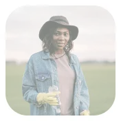 Woman wearing a denim jacket and black hat standing outdoors holding a glass of orange juice.