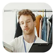 Young man with curly hair wearing a white shirt and a measuring tape around his neck, holding a clipboard and examining clothing on a rack.