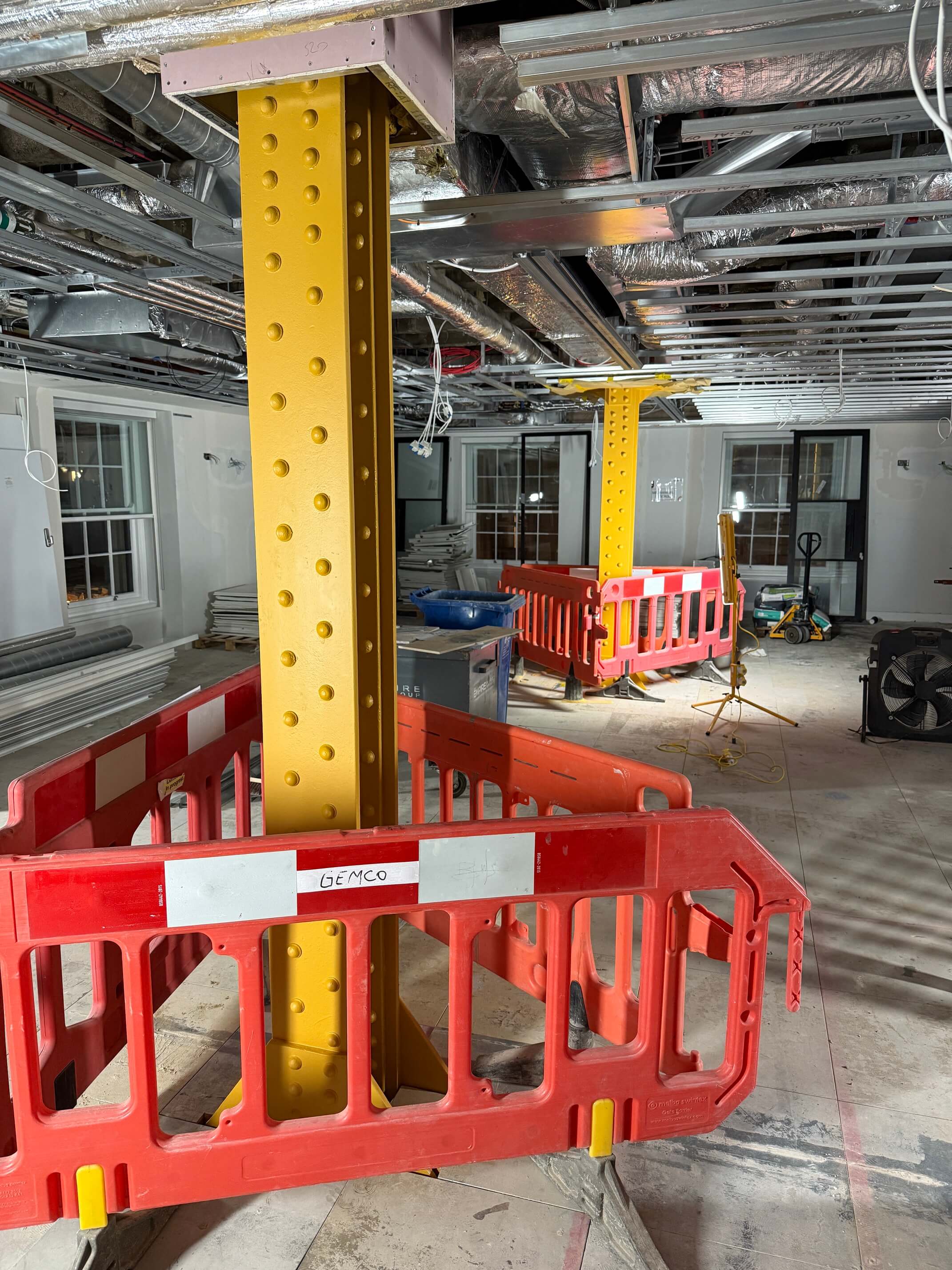 Indoor construction site with yellow steel support columns surrounded by red plastic safety barriers.