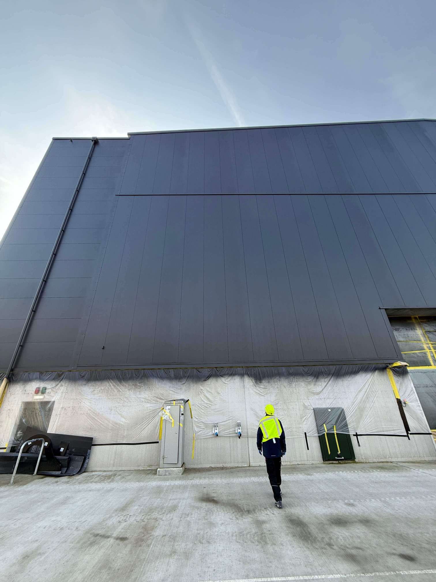 Person wearing high-visibility jacket walking toward a large dark gray industrial building with lower walls covered in protective plastic sheeting.