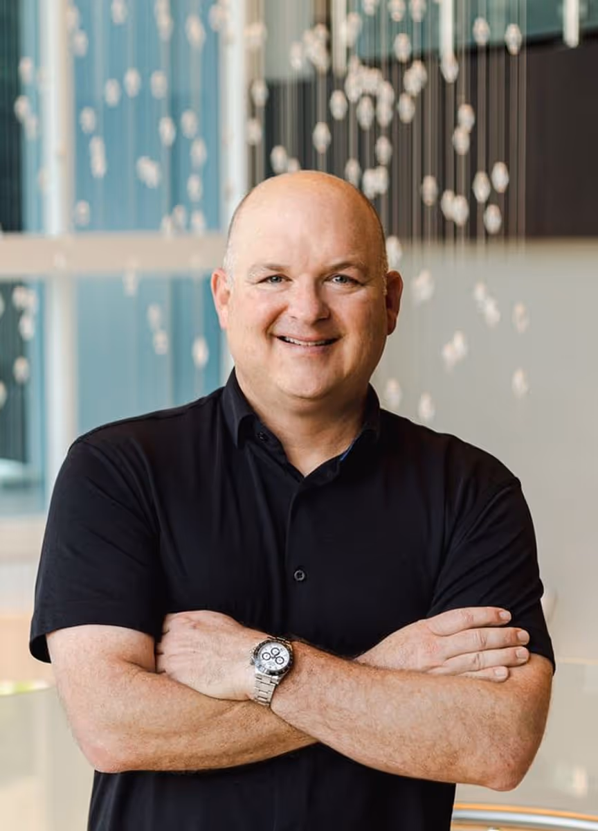 Smiling bald man in a black shirt with arms crossed, standing indoors with decorative hanging orbs in the background.