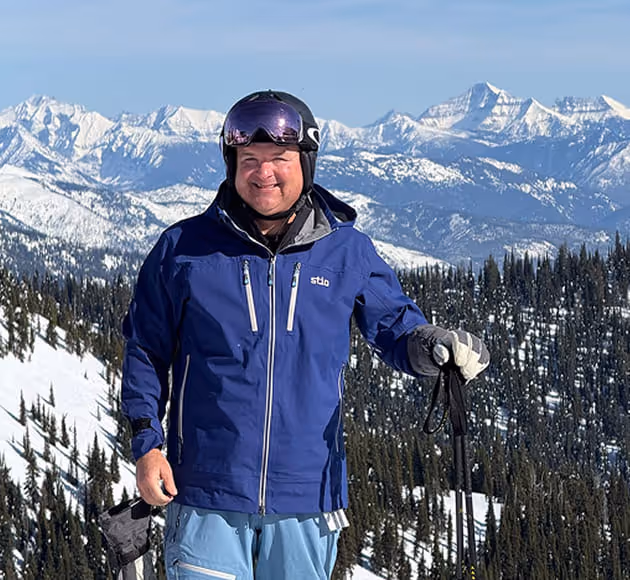 Smiling man in blue ski jacket and helmet standing on a snowy mountain with ski poles, with snow-covered forest and distant mountain peaks in the background.