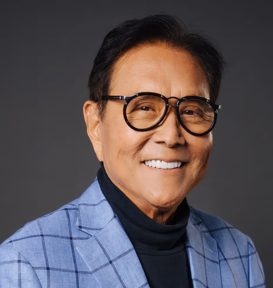 Smiling older man wearing large round glasses, a light blue checkered blazer, and a black turtleneck against a dark gray background.