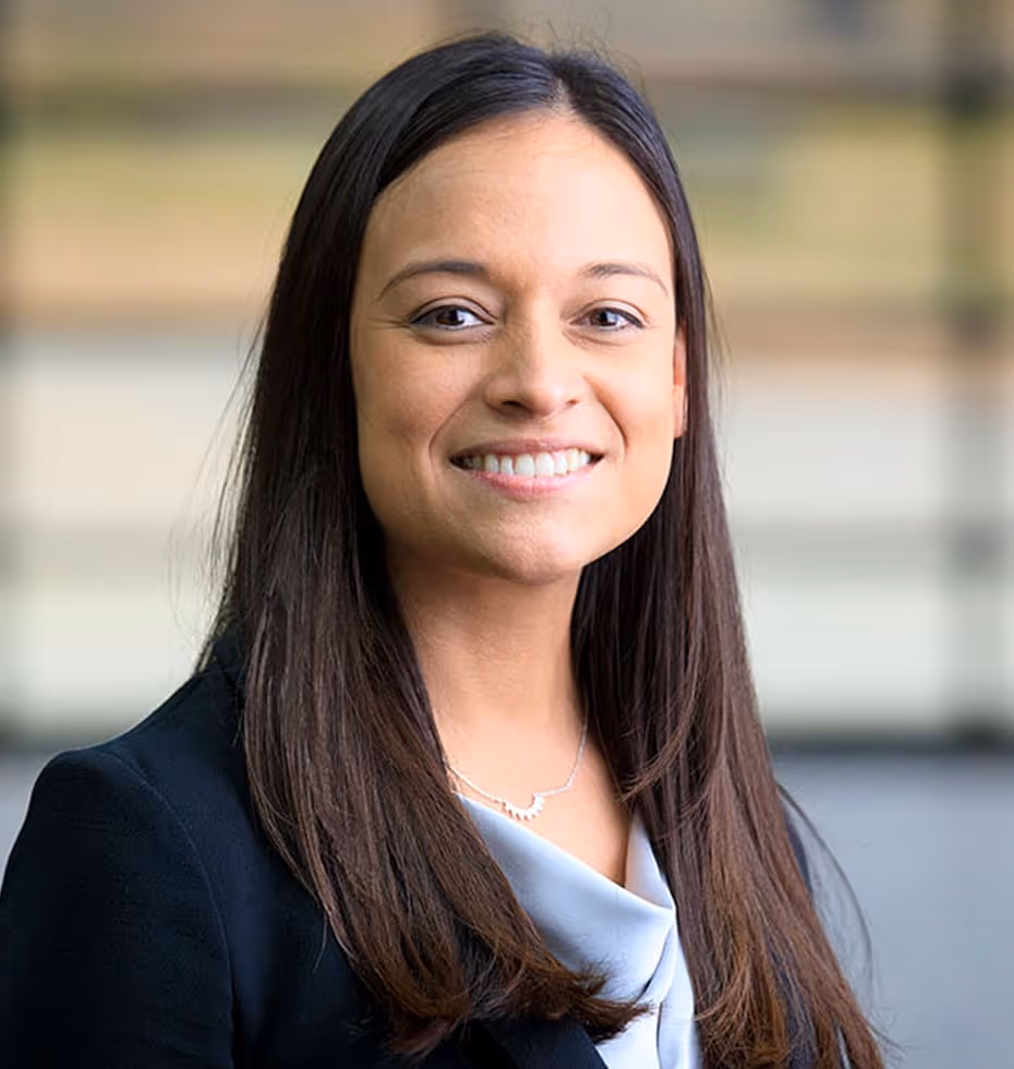 Smiling woman with long dark hair wearing a black blazer and light blouse in a blurred indoor setting.