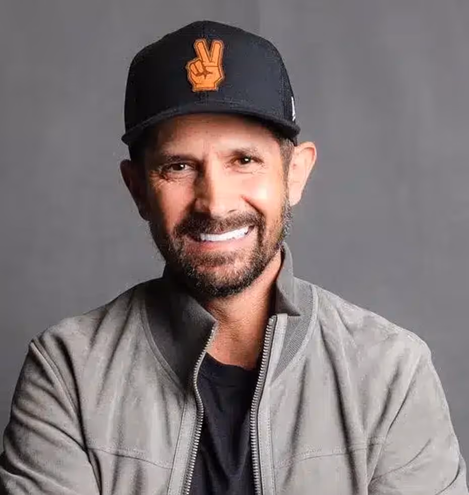 Smiling man with beard wearing a black cap with an orange peace hand symbol and a gray jacket against a gray background.