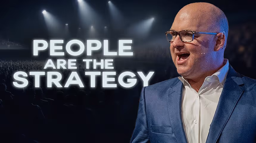 Bald man wearing glasses and a blue suit speaking on stage with illuminated text reading 'PEOPLE ARE THE STRATEGY' in the background.