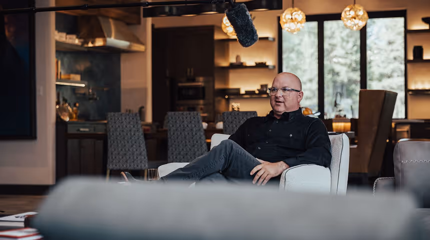 A bald man wearing glasses and a black shirt sits cross-legged in a white armchair in a modern, warmly lit room with dining chairs and large windows in the background.