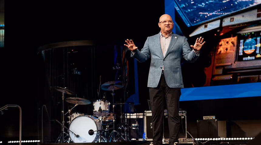 Man in a gray blazer speaking on stage with a drum set and a large screen behind him.