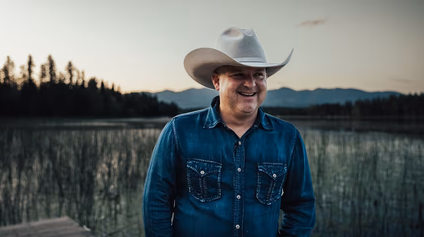 Smiling man wearing a cowboy hat and denim shirt standing by a lake with mountains and trees in the background at dusk.