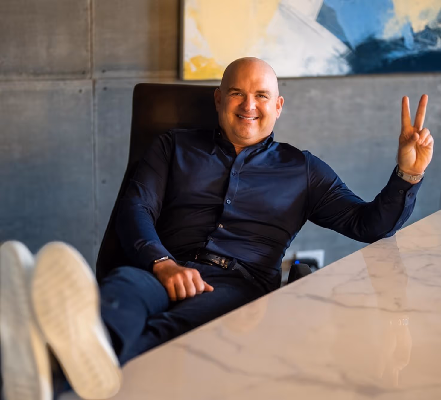 Smiling bald man in dark shirt sitting relaxed at a marble table with feet up and making a peace sign.