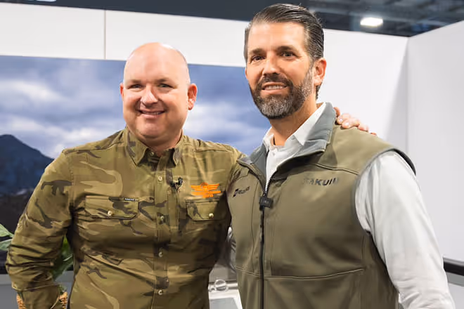 Two men posing together, one wearing a camouflage shirt and the other in a green vest over a white shirt.