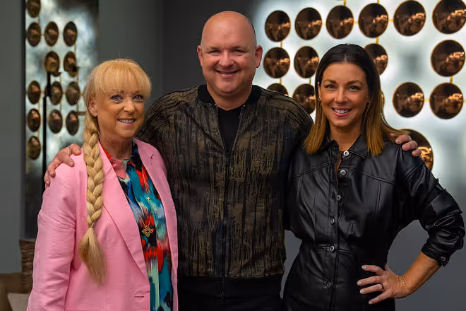 Three smiling adults posing together indoors with circular art on the wall behind them.