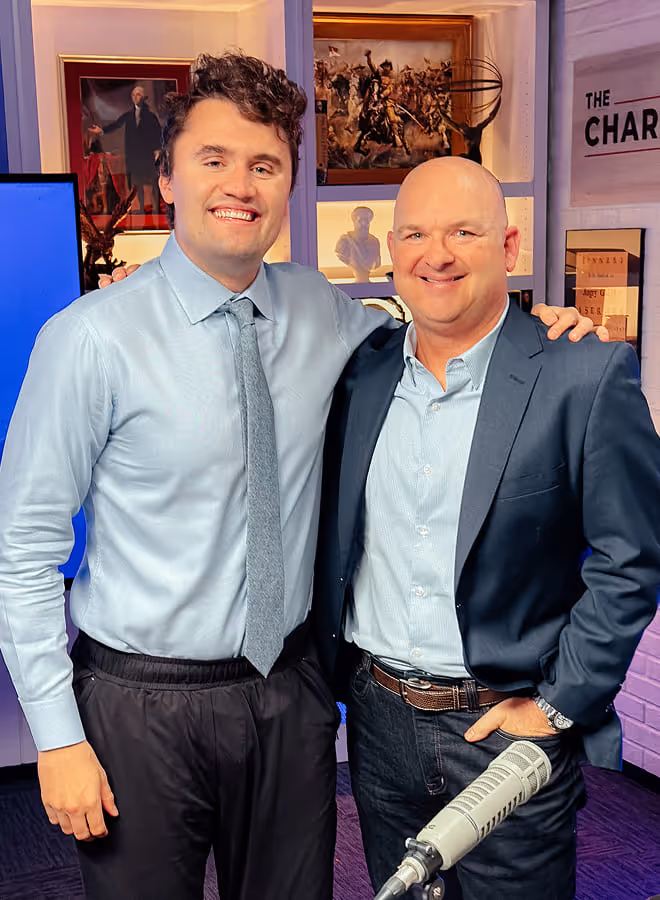 Two men smiling and posing in a studio with microphone; one man wears a blue shirt and tie, and the other wears a blue shirt with a navy blazer.