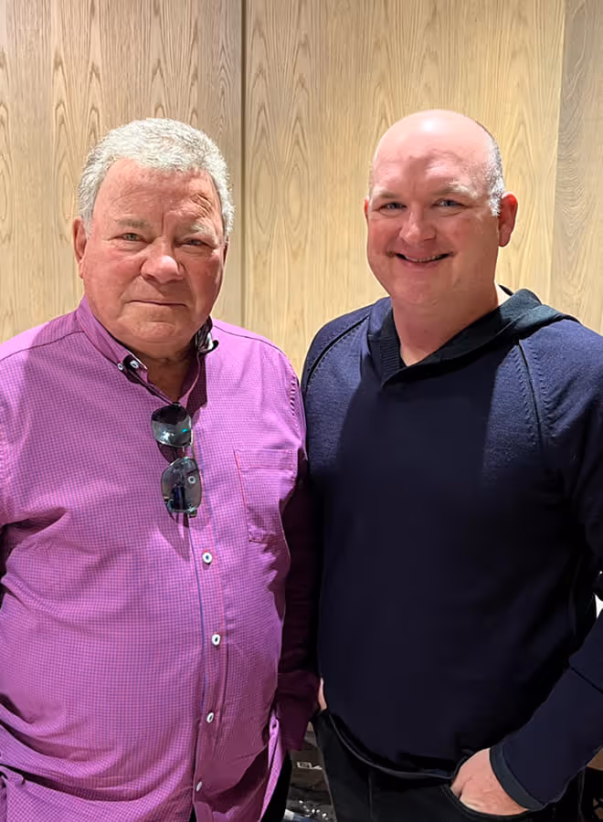 Two men standing indoors against a wooden panel wall, one wearing a purple checkered shirt with sunglasses hanging from the collar, the other in a black hoodie with hands in pockets.