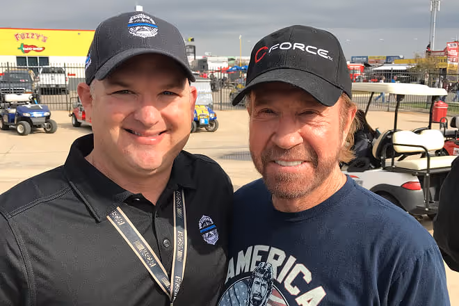 Two men smiling outdoors near golf carts, both wearing black hats and casual shirts.