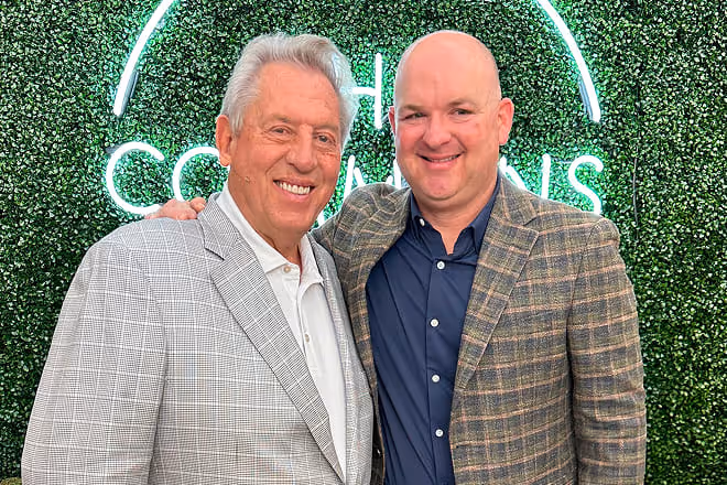 Two men smiling and posing together in front of a green leafy wall with neon text.