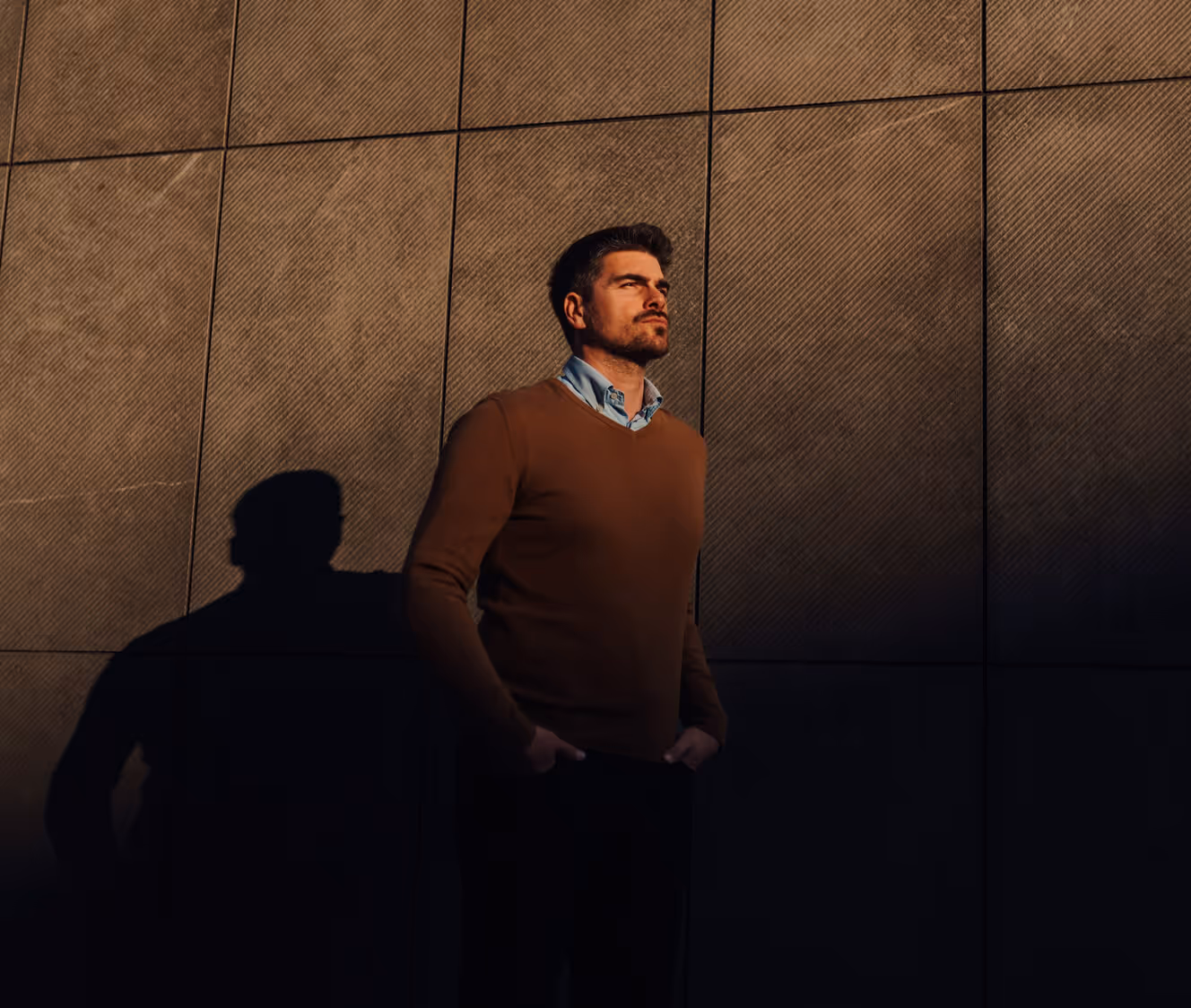 Man with hands in pockets wearing a brown sweater and blue shirt standing against a textured wall with his shadow visible.