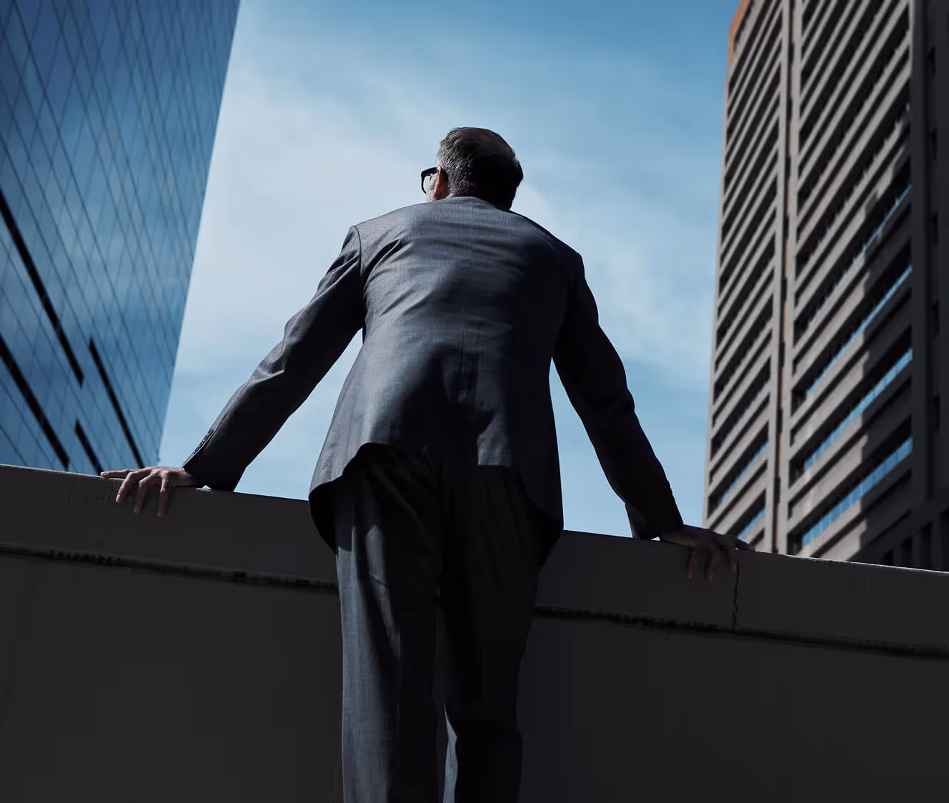 Man in a suit leaning on a ledge, looking up between two tall buildings under a blue sky.