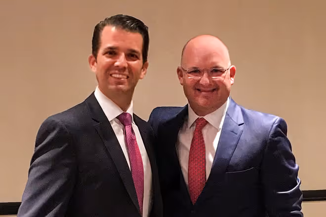 Two men in dark suits and red ties smiling and posing together against a plain beige background.