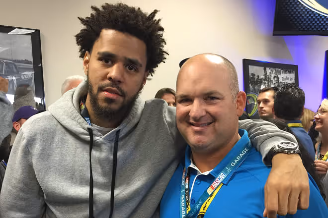Two men posing with arms around shoulders at an indoor event with people in the background.