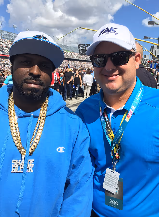 Two men in blue clothing posing at a crowded outdoor event; one wears a blue hoodie with gold chains and a white cap, the other a blue shirt, sunglasses, and a white cap with a lanyard and credentials.