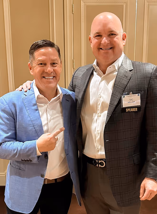 Two men smiling indoors, one wearing a blue blazer pointing at the other man who wears a dark checked blazer and a speaker name tag.