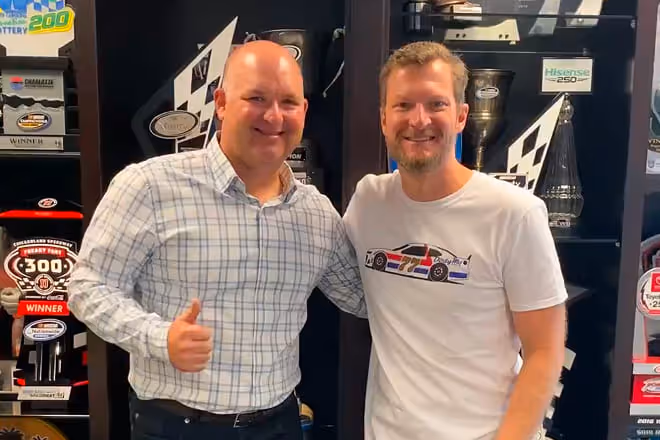 Two men smiling and posing together in front of a display case with racing trophies and memorabilia.