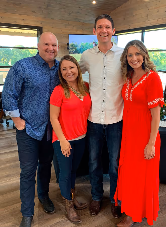 Four smiling adults standing indoors against wooden walls and large windows, two men and two women dressed casually.