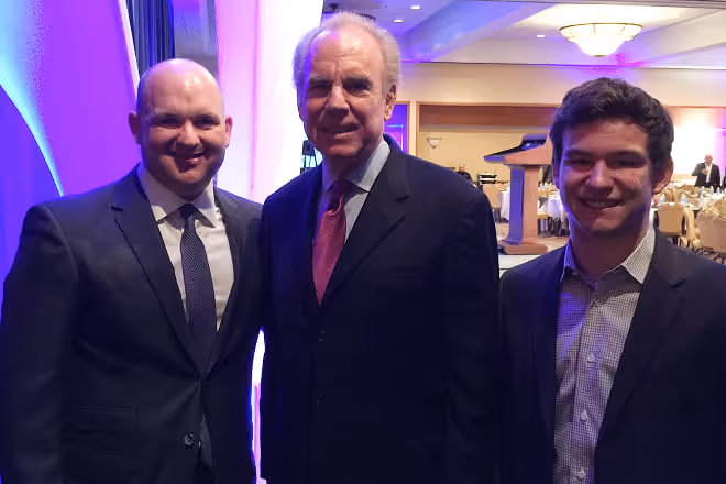 Three men in suits standing and smiling at an indoor event with round tables and a podium in the background.