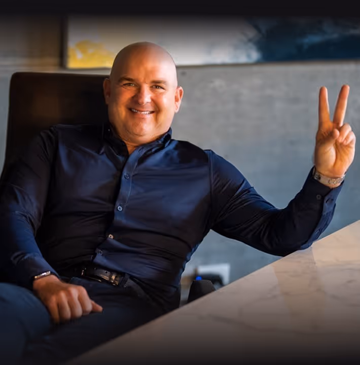 Smiling bald man in a dark shirt sitting at a table and making a peace sign with his right hand.