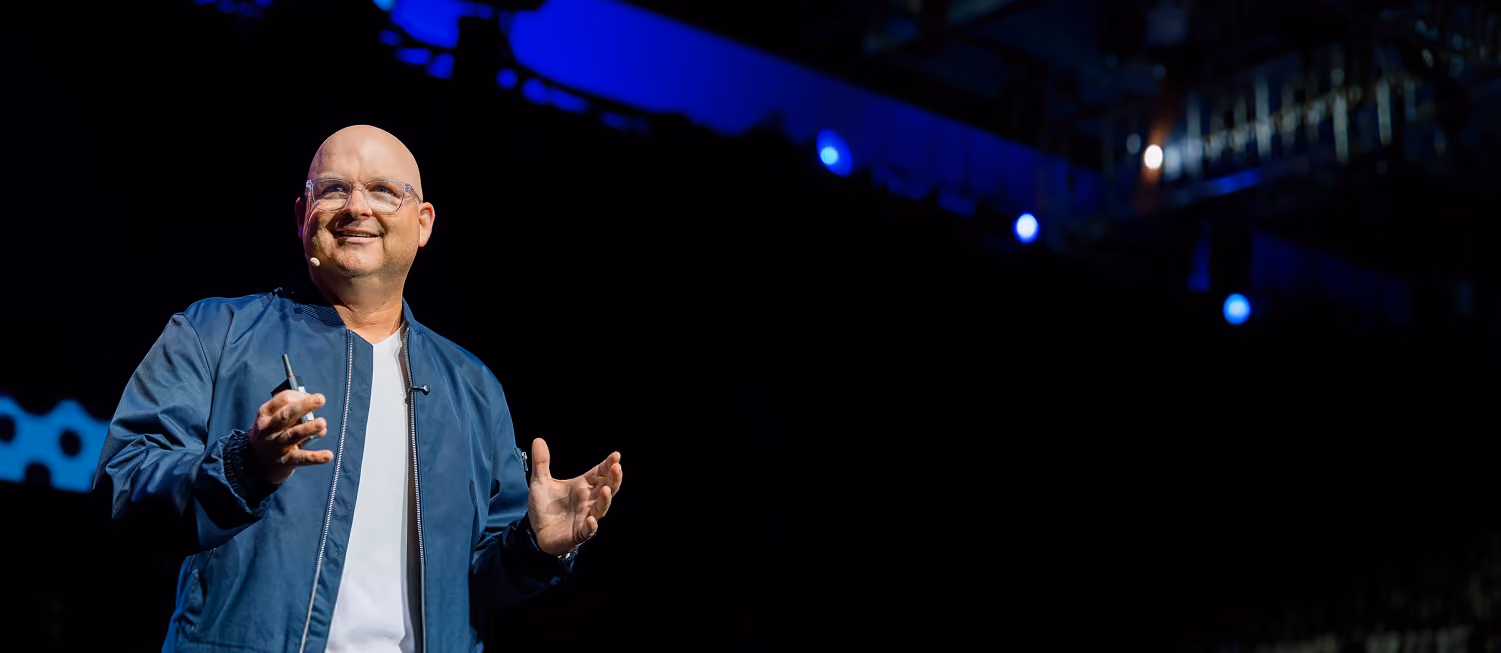 Bald man wearing glasses and blue jacket speaking on stage with a microphone headset and hand gestures.