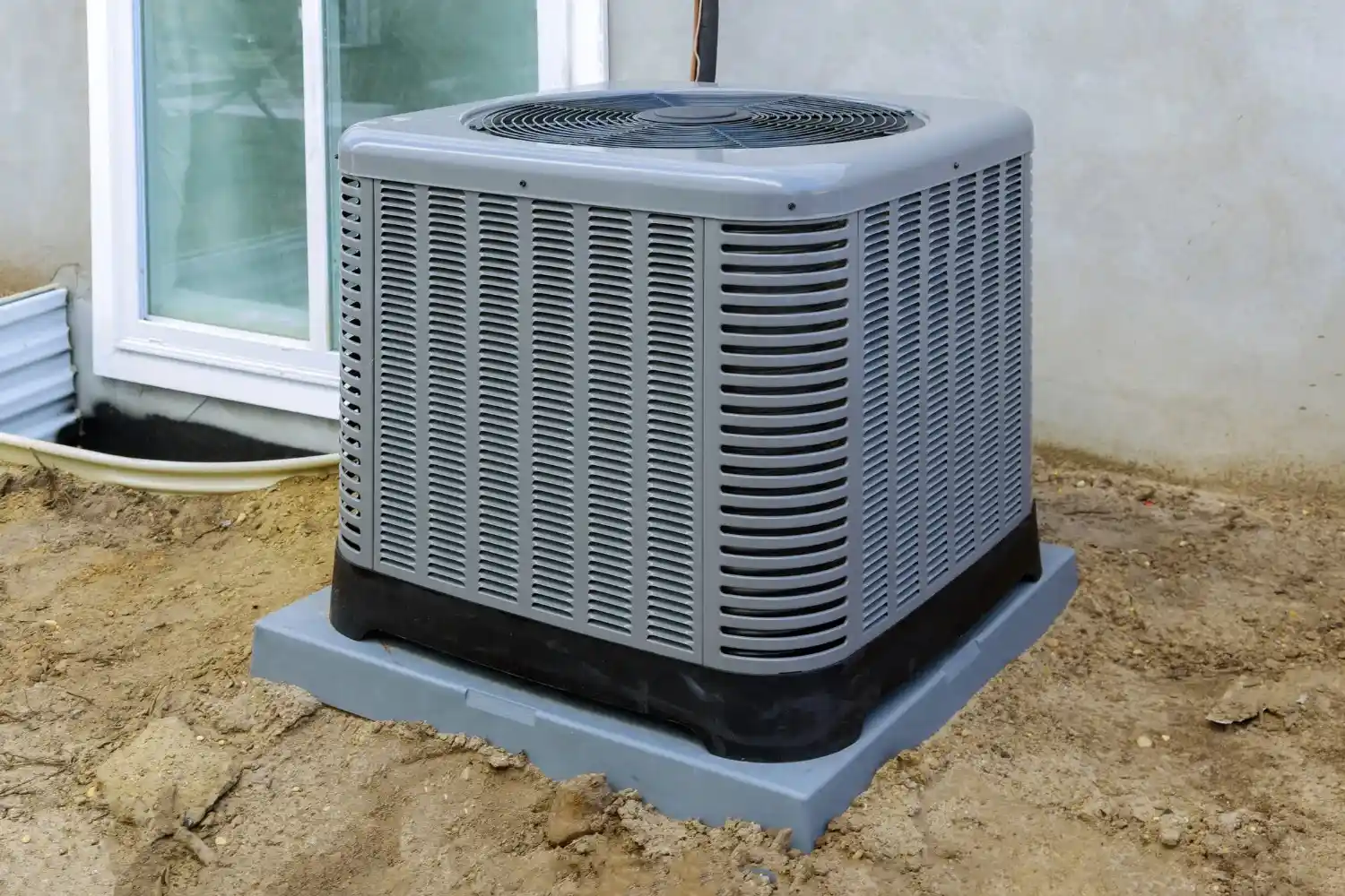A single grey heat pump condenser unit with a fan on top, sitting on a plastic or composite base on a patch of dirt next to a house foundation with a small window