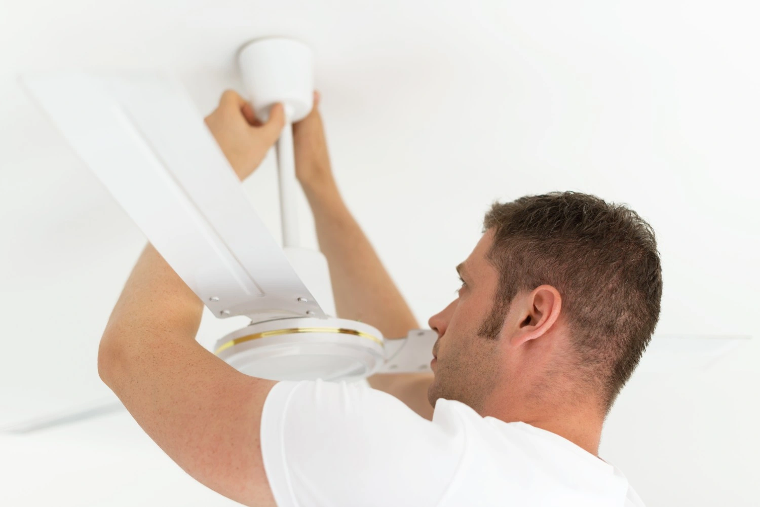 A man in a white t-shirt installing or repairing a white ceiling fan against a white ceiling.