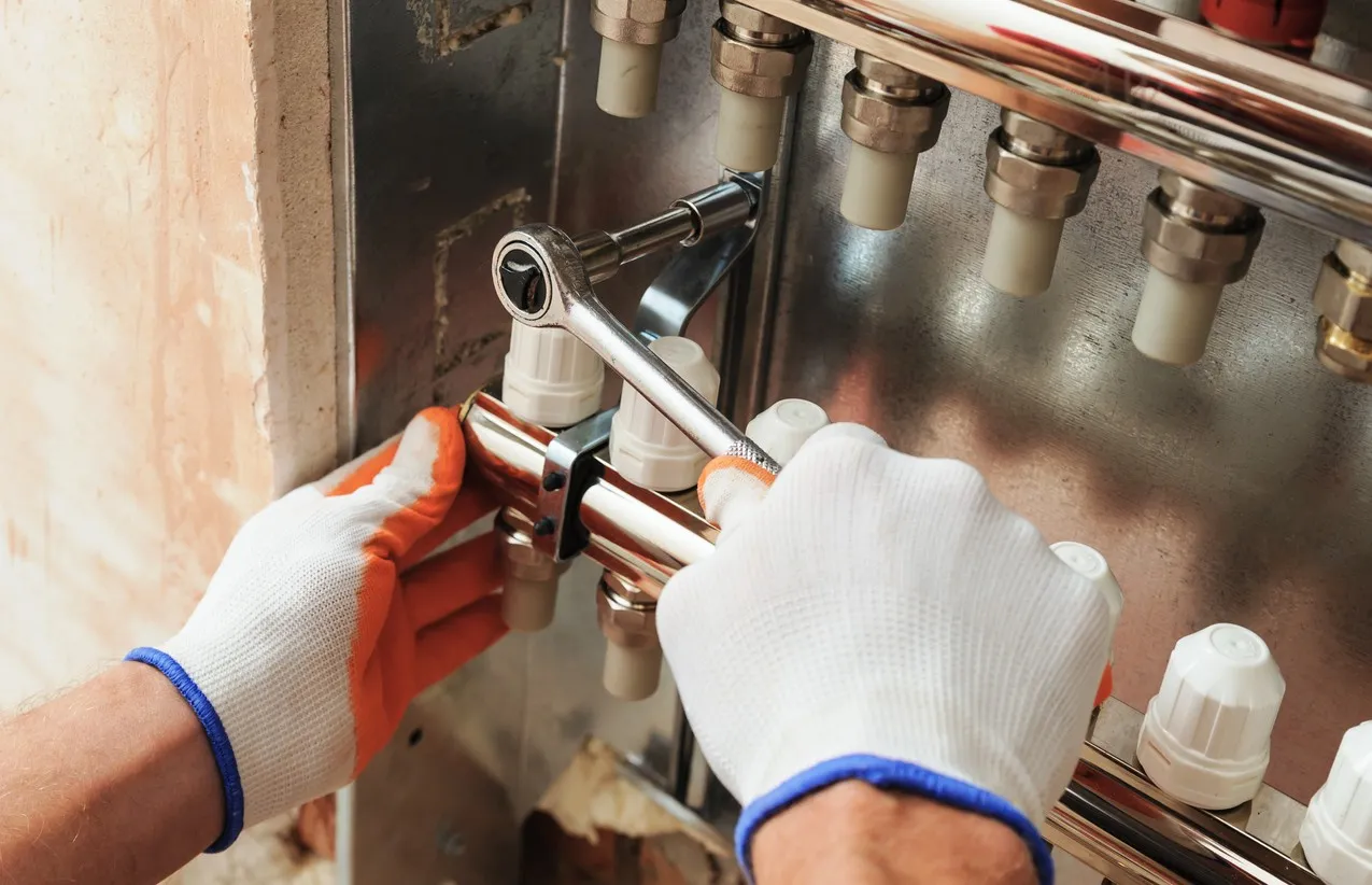  A close-up shot shows a pair of hands wearing white work gloves with orange and blue trim, actively working on a heating or plumbing manifold. The right hand is using a ratchet and socket to tighten a metallic fitting on a chrome pipe, which is connected to multiple white plastic pipe inlets. The left hand is steadying the manifold. The background shows a raw wall recess where the system is mounted.