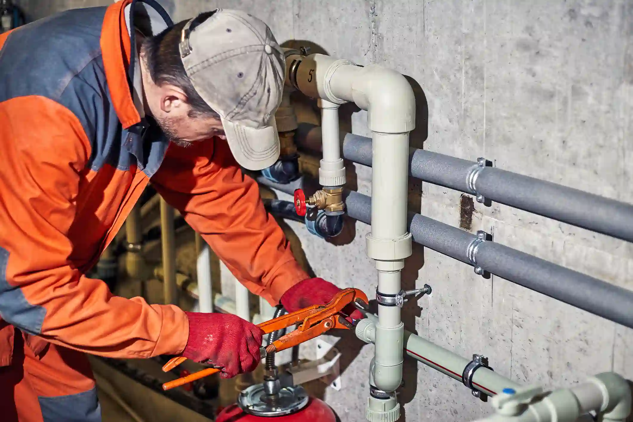 A technician in orange overalls and red gloves uses a large orange pipe wrench to service beige piping against a concrete wall in a mechanical room.