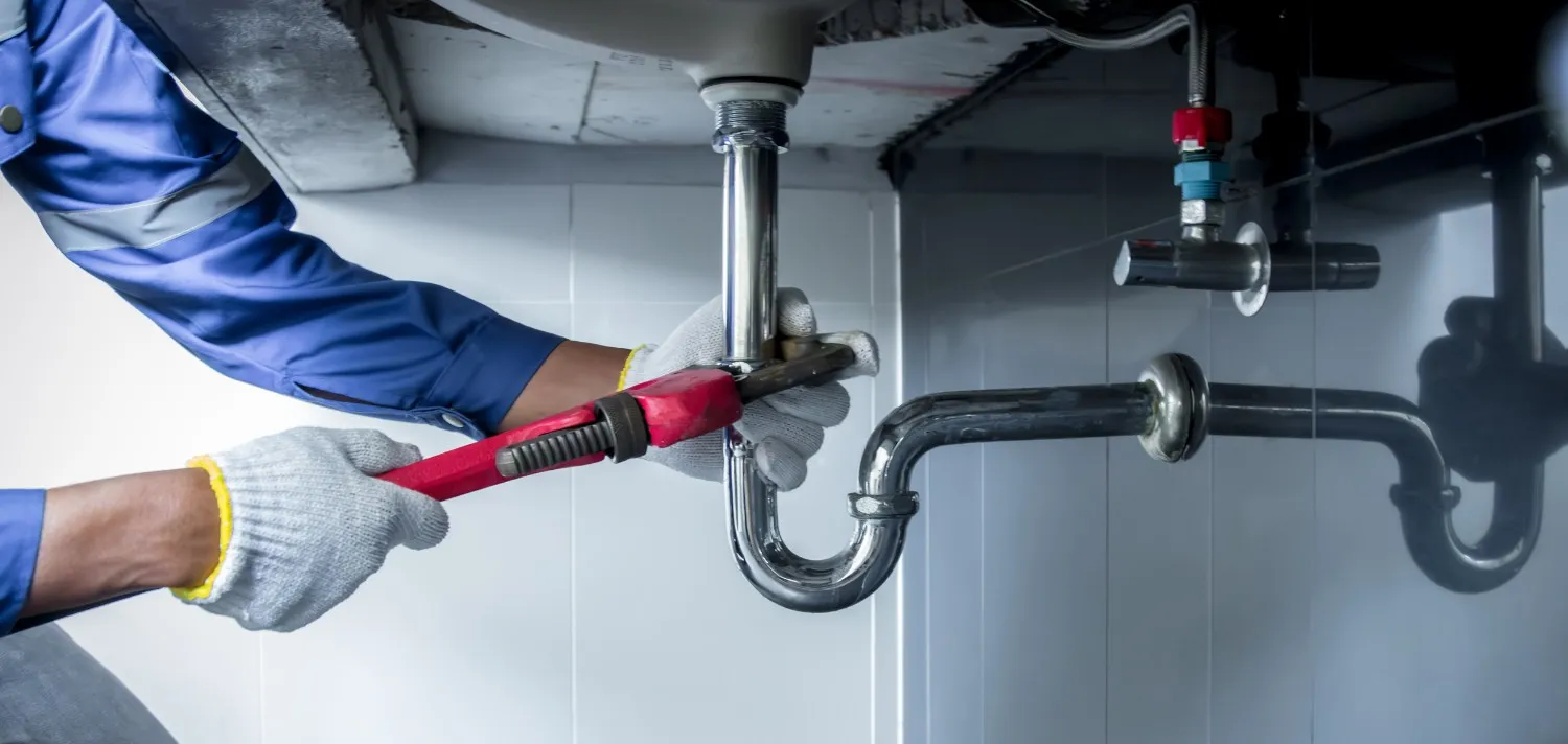 A plumber wearing white gloves and a blue uniform uses a large red pipe wrench to tighten or repair a chrome-plated P-trap drainage pipe underneath a white commercial sink.