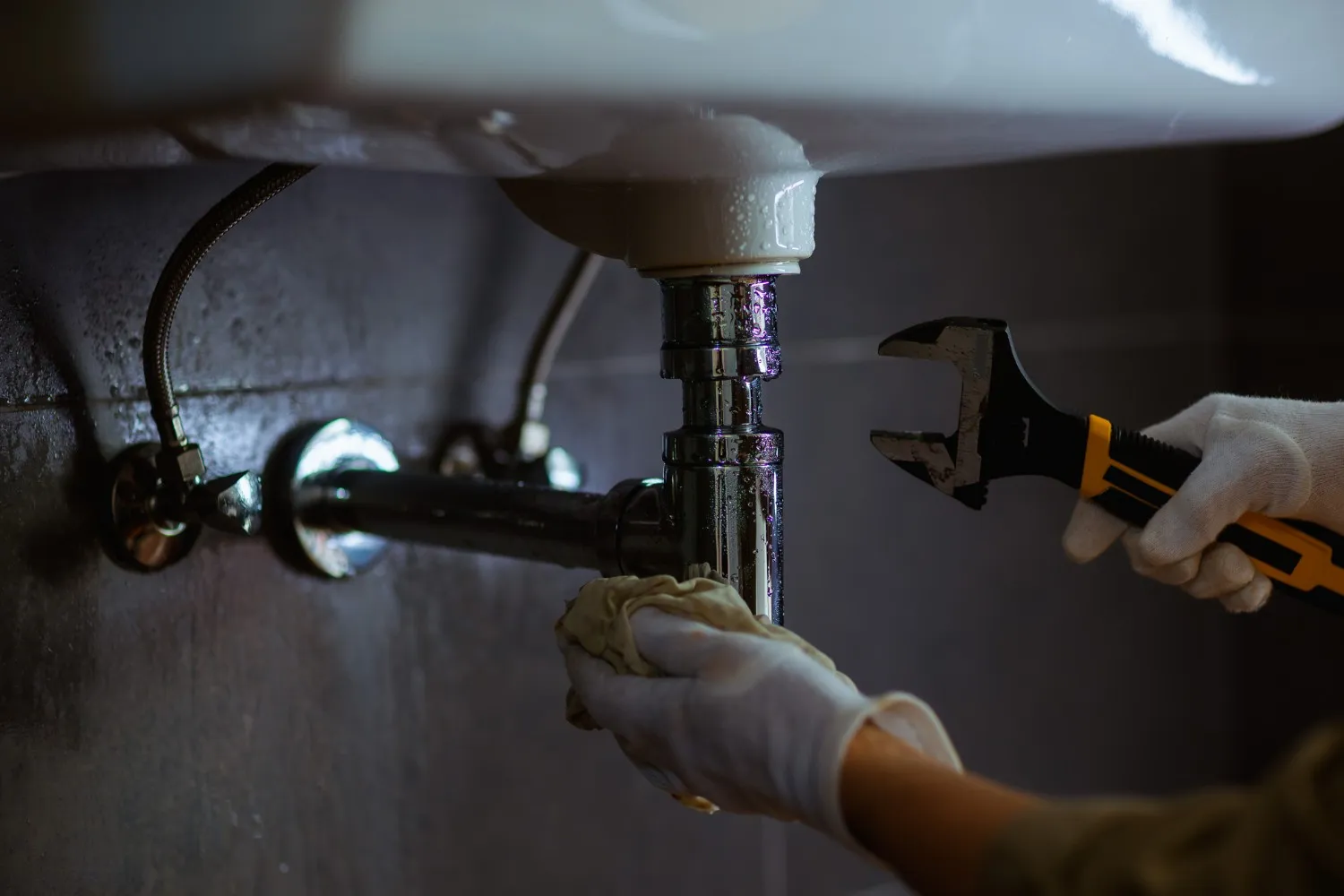 A plumber wearing white gloves uses an adjustable wrench and a cloth to repair a chrome P-trap drainage pipe under a bathroom sink. The scene shows a close-up of the plumbing work against a dark tiled wall.