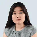 Portrait of a young woman with long dark hair wearing a gray top against a white background.